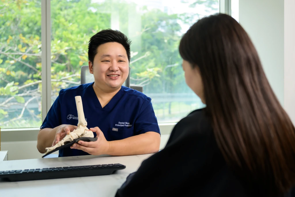 A podiatrist in navy scrubs uses a skeletal foot and ankle model to explain anatomy to a patient deciding whether to see a podiatrist or orthopedist for foot pain.