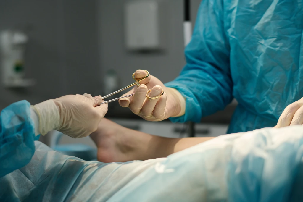 Gowned theatre staff passing a surgical instrument over a draped bare foot in an operating theatre, the setting behind questions about Morton's neuroma surgery side effects.