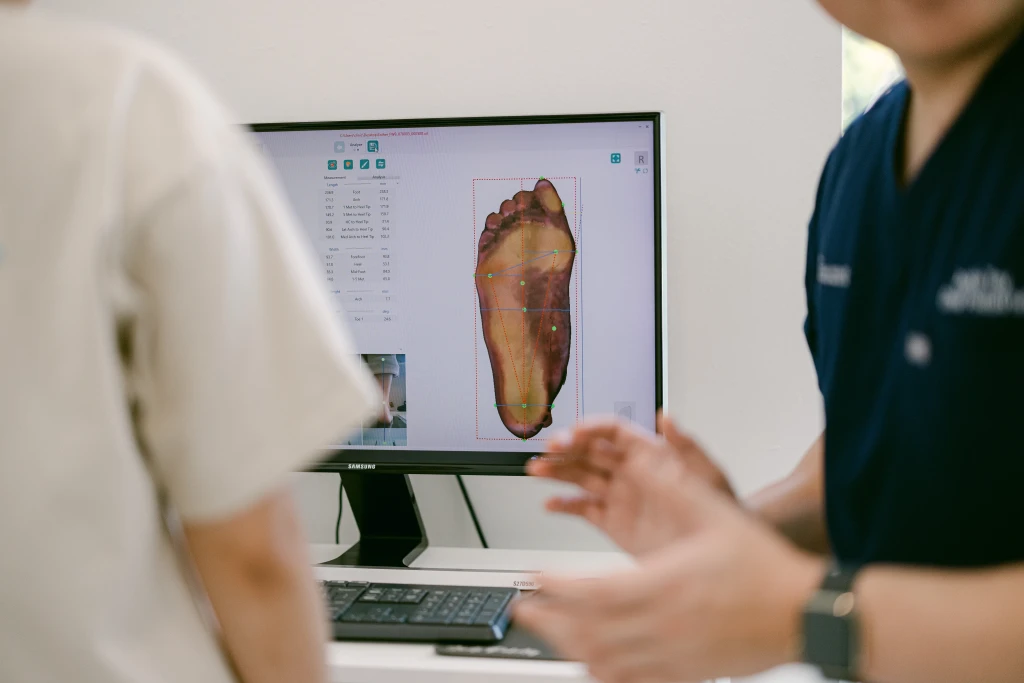 A clinician in navy scrubs discussing an on-screen digital scan of a patient's sole with plotted arch measurements, the analysis used to identify foot arch types