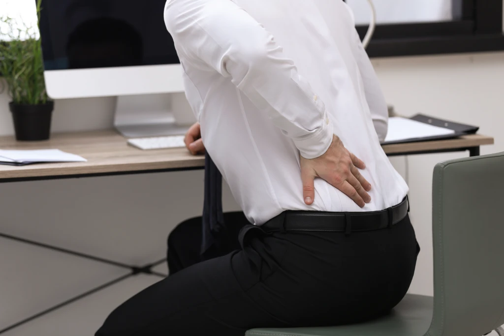 A person seated at an office desk pressing one hand to their lower back, the pelvic and lower back pain pattern described in malalignment syndrome.