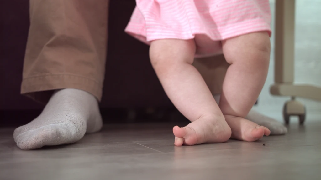 Close-up of a baby's bare feet taking steps on a tiled floor beside an adult's socked foot, the walking stage when out toeing in children is first noticed
