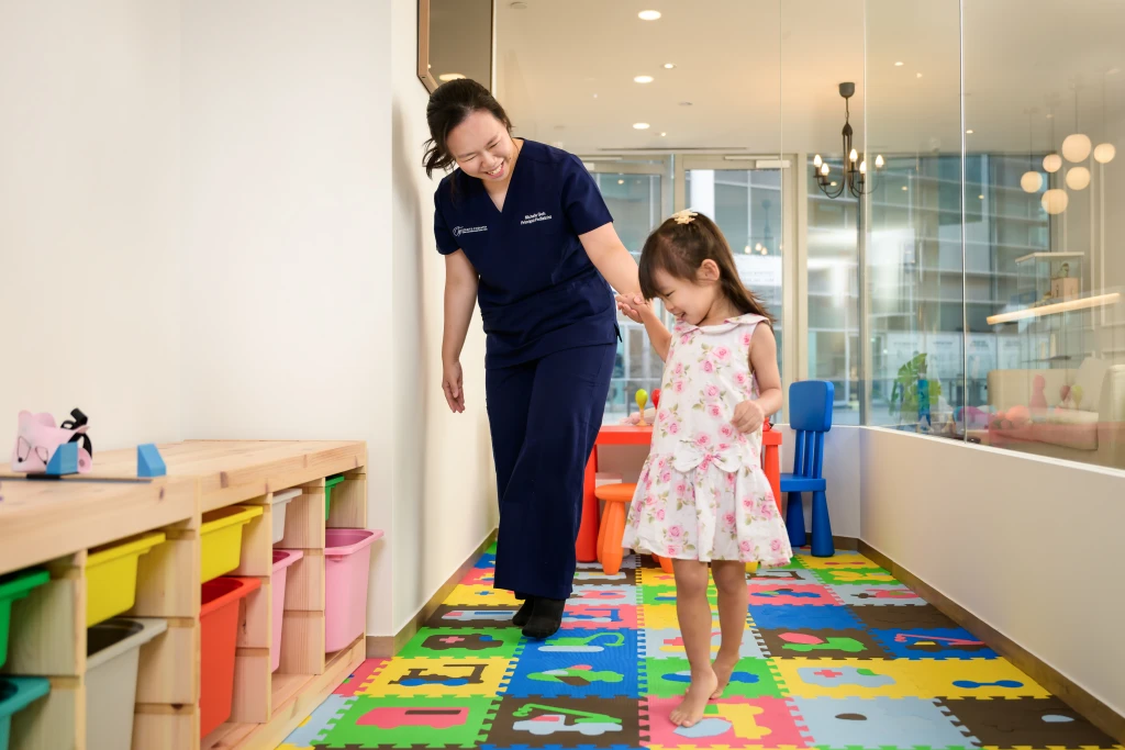 A podiatrist in navy scrubs walking hand in hand with a young child across a colourful play mat in a clinic room set up to assess postural issues in children.