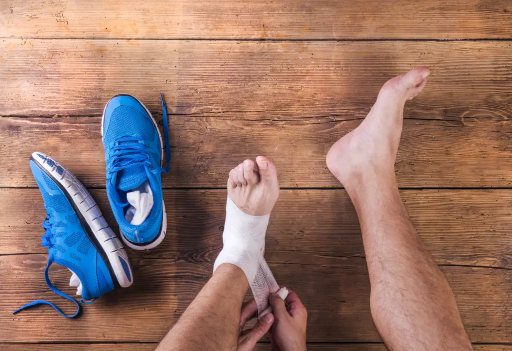 An overhead view of a person bandaging their own ankle on a wooden floor beside blue running shoes, immediate self-care for the most common foot and ankle injuries