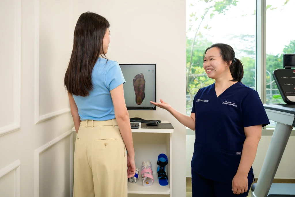 A podiatrist in navy scrubs gestures to a foot scan image on a clinic monitor while a standing patient looks on, part of what a podiatrist does on a first visit.