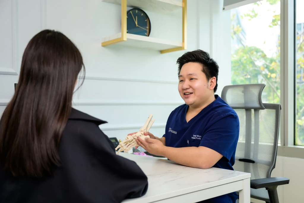 A podiatrist in navy scrubs uses a foot skeleton model to explain anatomy to a patient across a clinic desk, the clinical role behind the question is a podiatrist a doctor