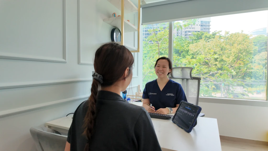 A podiatrist in navy scrubs speaking with a patient across a clinic desk beside a tablet of results, the discussion that is part of what to expect at a podiatry appointment.