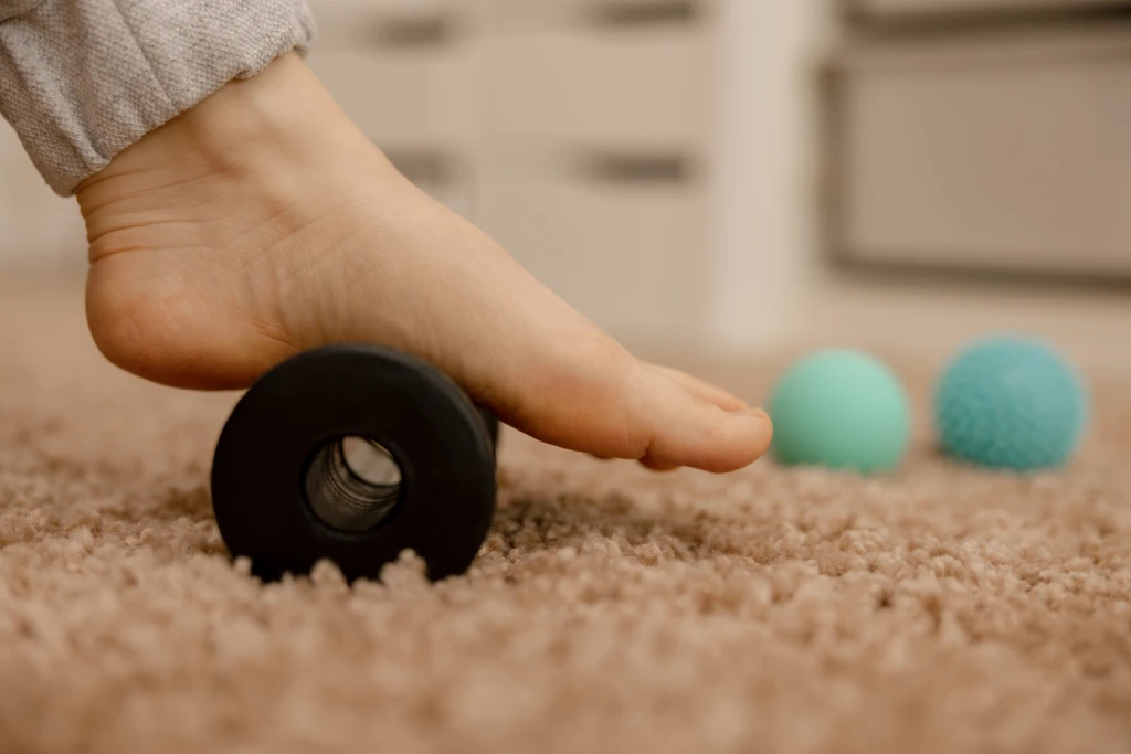 A bare foot rolls its arch over a black massage roller on carpet beside two massage balls, self-care tools tried when plantar fasciitis is not getting better with stretches