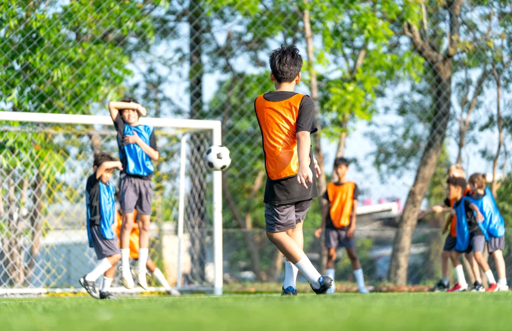 Young footballers in orange and blue training bibs playing on an artificial turf pitch, the surface and pace behind many common football injuries.