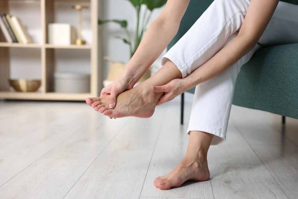 A person sitting on a sofa at home clasping one bare foot with both hands, the persistent foot pain behind questions about plantar plate tear recovery time.
