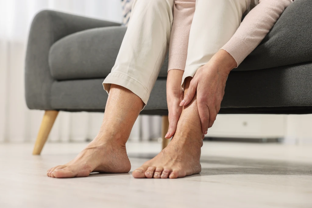 A person sitting on a sofa at home reaching down to hold their lower leg above two bare feet resting on the floor, the moment behind asking why does my foot hurt more after resting