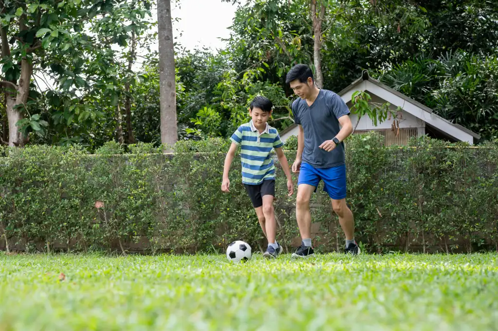 An adult and a child in trainers playing football on a grass lawn, the repeated kicking and stopping that brings on toenail pain after football.