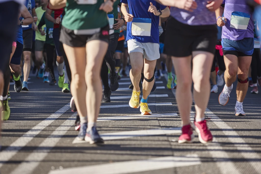 A tight pack of race runners photographed from the waist down striding along a sunlit marked road, the repeated loading that raises the question of how to avoid running injuries.