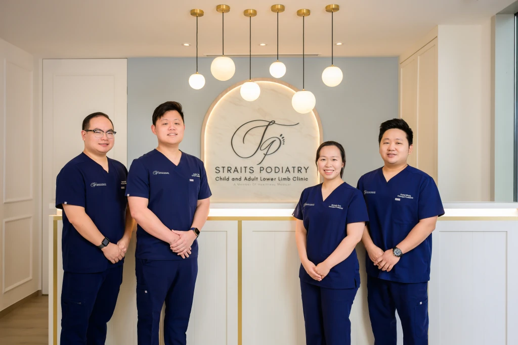 Four podiatrists in navy scrubs stand at a clinic reception desk beneath the Straits Podiatry sign, the team and setting to weigh up in how to choose a podiatry clinic