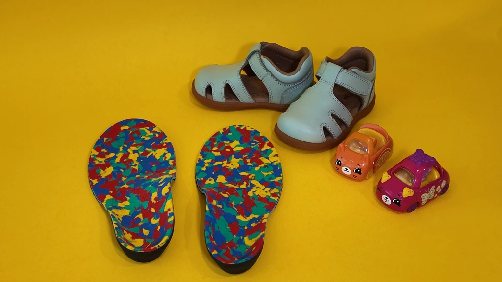 A pair of children's closed-toe sandals beside two colourful child-sized orthotic insoles and toy cars, the supportive build behind podiatrist recommended shoes for kids.