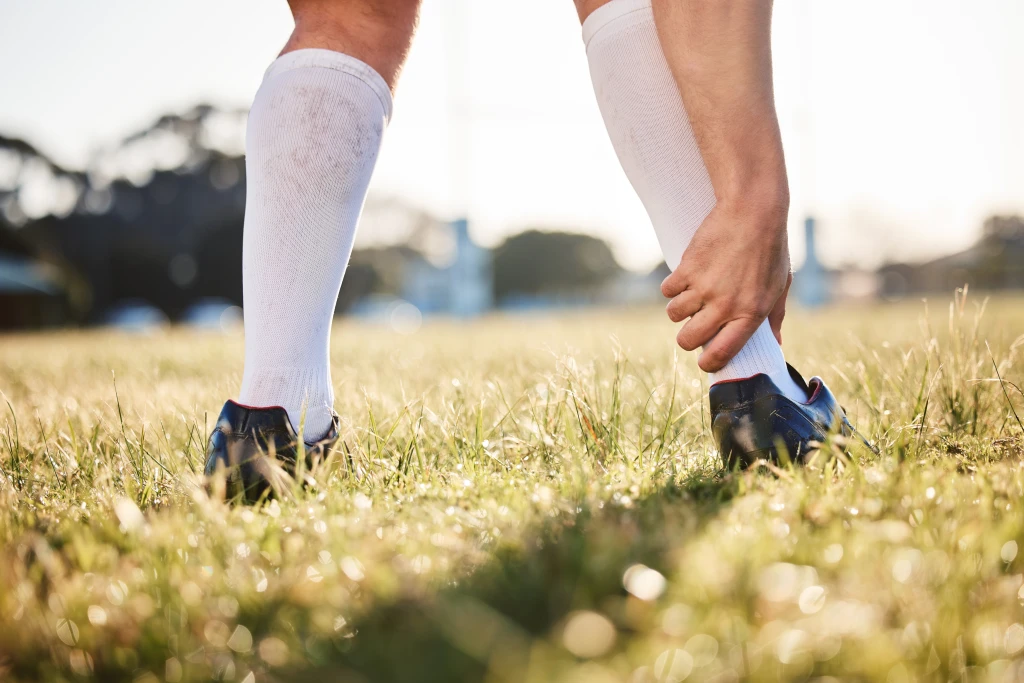 A footballer in boots and white socks on a grass pitch reaching down to grip the back of the heel, where an Achilles tendon tear is felt.