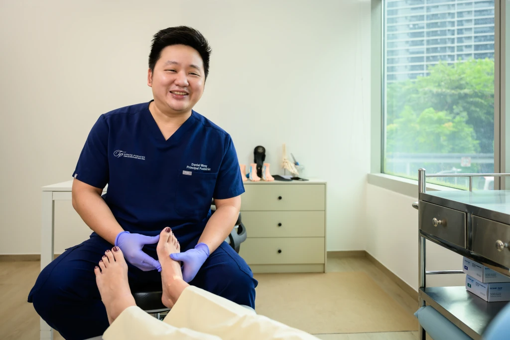 A podiatrist in navy scrubs and gloves cradles a patient's bare foot in a bright consultation room, the hands-on assessment that makes up a foot health screening in Singapore.