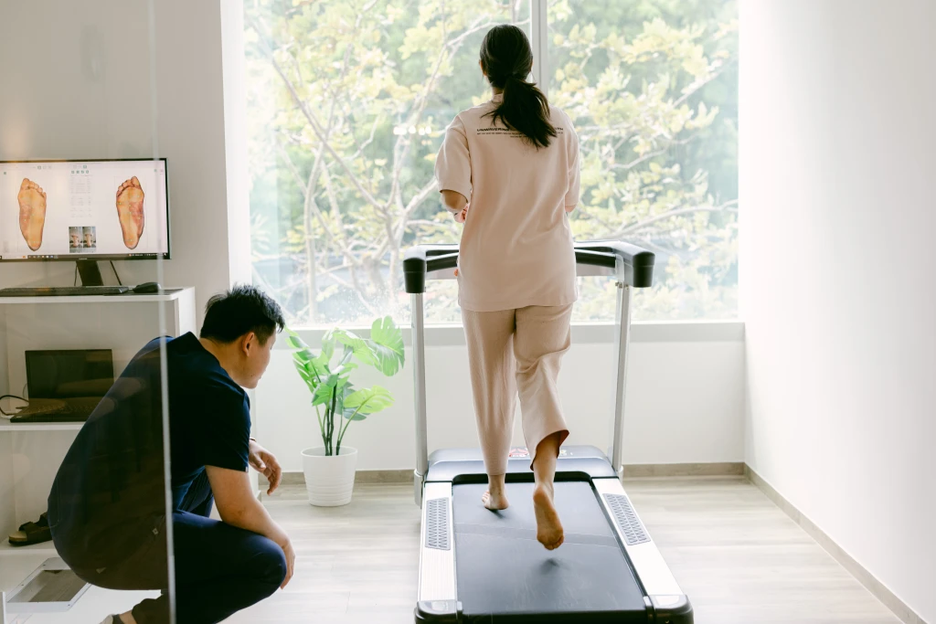 A clinician crouches to watch a patient walk barefoot on a clinic treadmill with foot scans on the monitor, the gait analysis behind podiatrist vs physiotherapist comparisons.