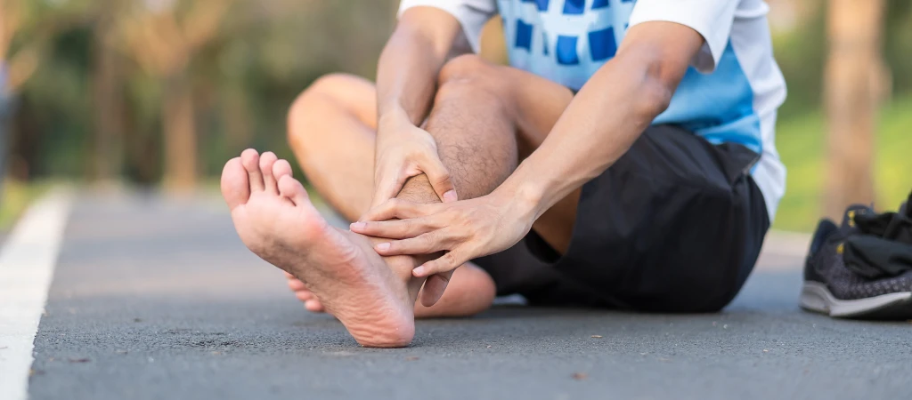 A person in sports kit sits on a park path gripping the heel and ankle of one bare foot, the lingering pain that signals when to see a podiatrist