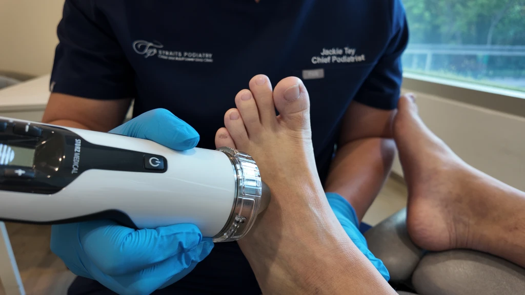 A podiatrist in navy uniform presses a handheld shockwave applicator to the top of a patient's bare foot, one way pain on top of foot is managed