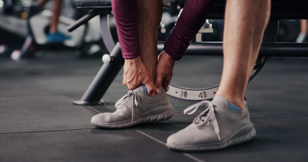A person in a gym bends to grip the outer ankle above a grey trainer, the guarding grasp typical when an ankle feels unstable after sprain