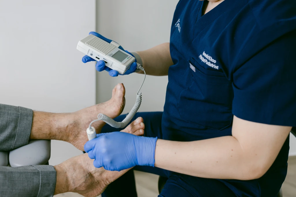 A podiatrist in gloves holding a handheld Doppler probe against a patient's toe to check blood flow, part of assessing how to tell if a diabetic foot is infected.
