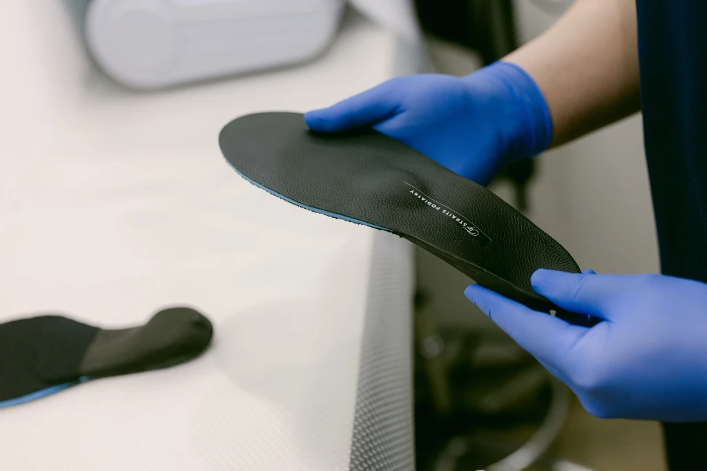 Gloved hands holding a black custom foot orthotic labelled Straits Podiatry beside a second shell, a device used when managing malalignment syndrome.