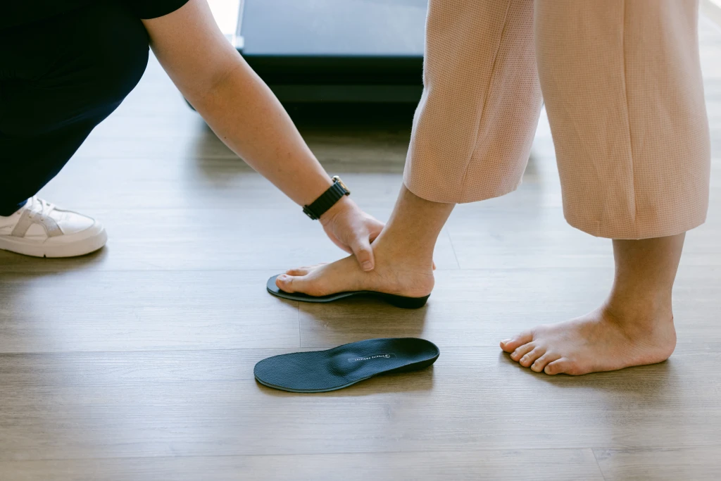 A podiatrist kneeling to steady a patient's bare foot on a black custom orthotic beside a second orthotic, the offloading that influences how long plantar fasciitis takes to heal.