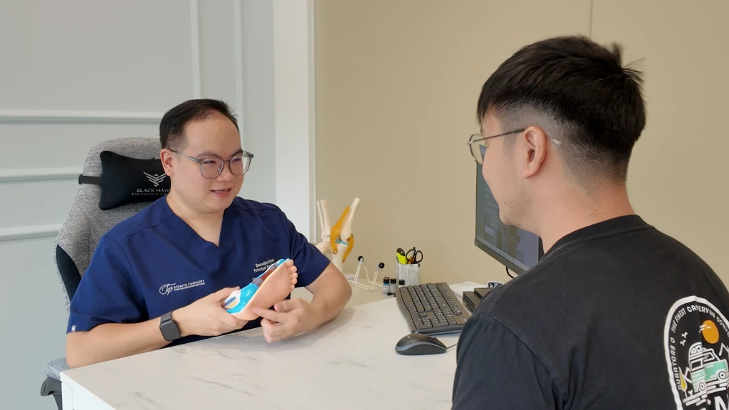 A podiatrist in navy scrubs holds a foot model across a desk to explain findings to a seated patient, showing what a podiatrist does for heel pain at a consultation