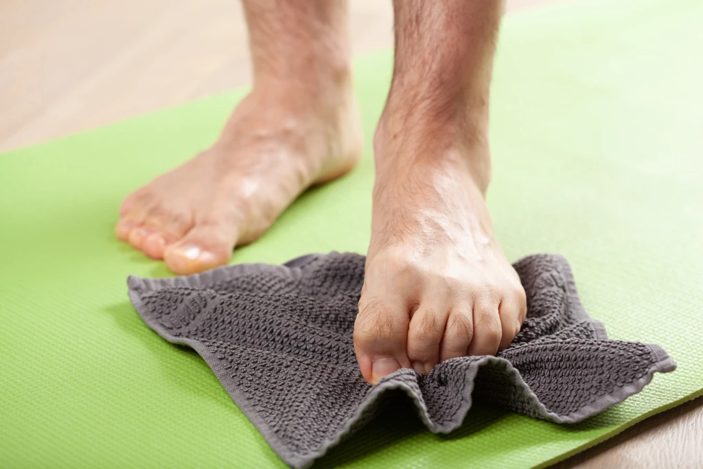 Bare feet on a green exercise mat with the toes of one foot scrunching a grey towel, the towel curl showing how to do plantar fasciitis exercises correctly.