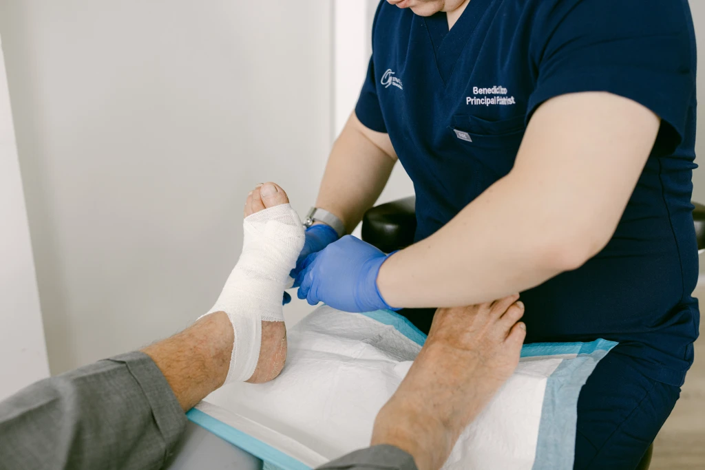 A podiatrist in navy scrubs and blue gloves wrapping a bandage around a patient's foot and ankle, the wound care used to protect tissue at risk of diabetic gangrene.