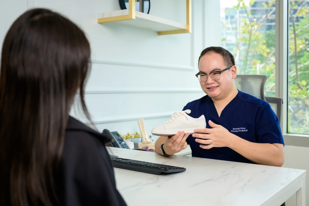 A podiatrist holds up a white lace-up sneaker to show a patient across a marble consultation desk, footwear review being one part of a foot health screening in Singapore.