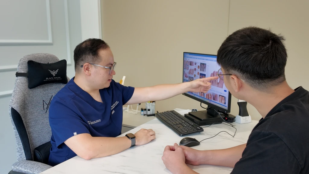 A podiatrist points to toenail photographs on a monitor while explaining them to a seated patient, the kind of consultation that covers how to prevent fungal nail infection.