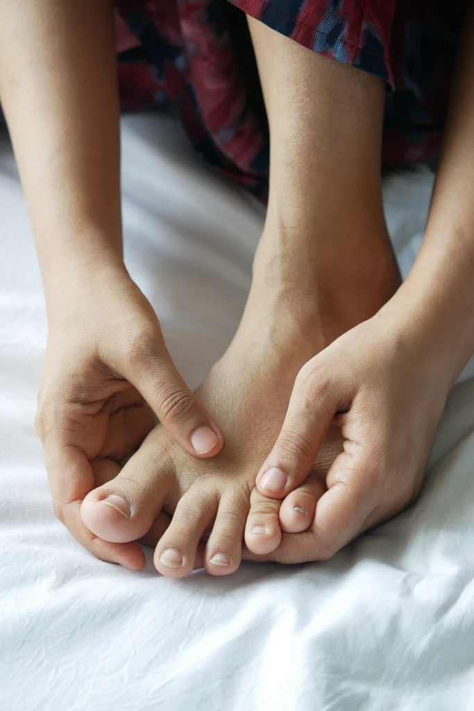 Close-up of a person seated on a bed squeezing the toes and forefoot of one bare foot, the forefoot area involved when weighing Morton's neuroma surgery side effects.