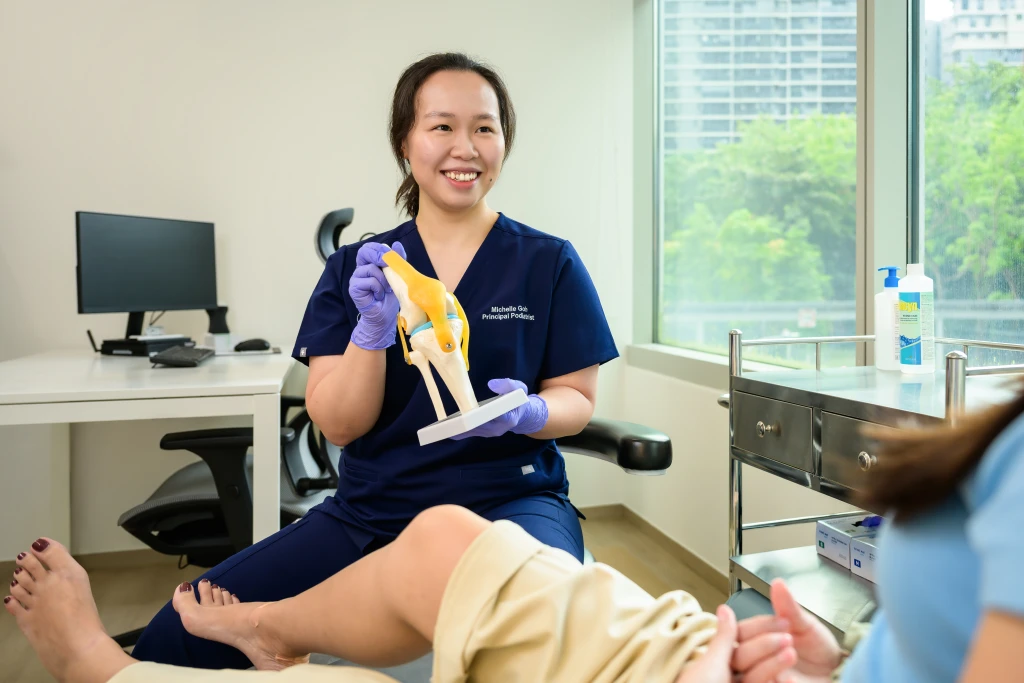 A podiatrist in a clinic room holding an anatomical knee joint model while speaking with a seated patient, the lower limb check-up encouraged for International Podiatry Day.