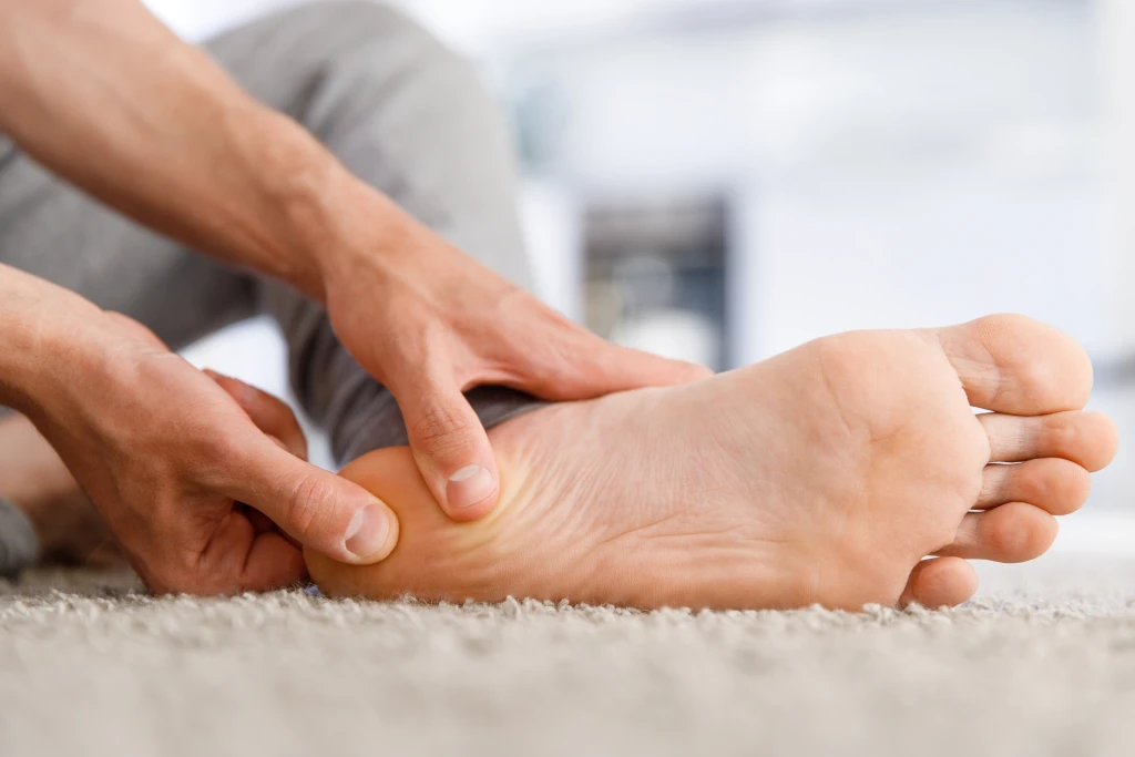 A person sitting on the floor pressing both thumbs into the arch of a bare foot, an orange glow marking the sore area, illustrating why does my foot hurt more after resting
