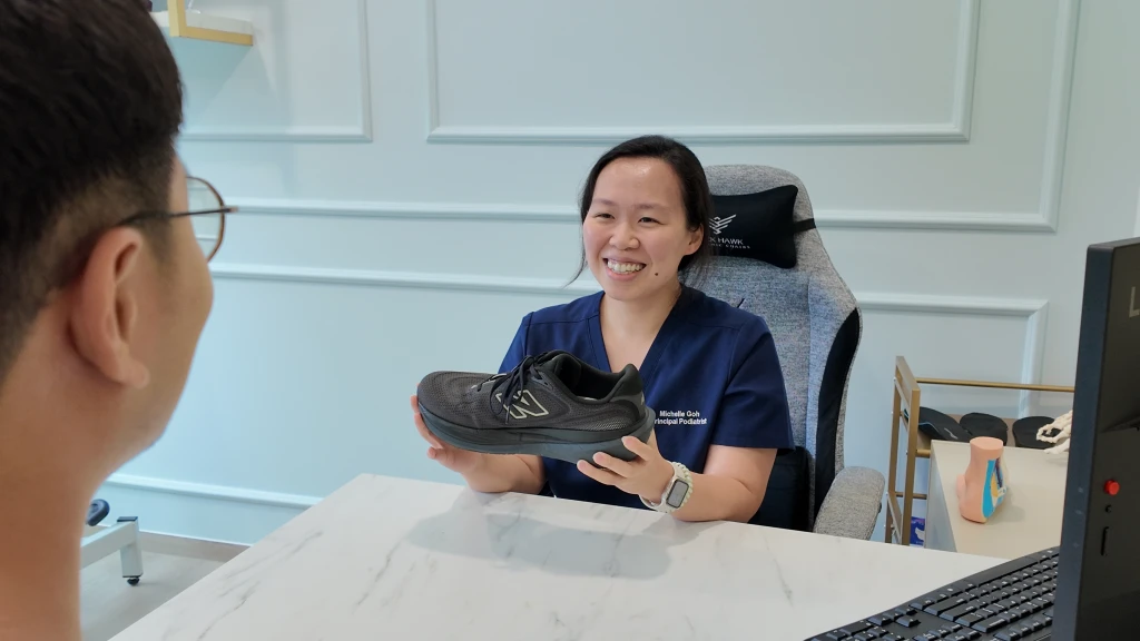 A podiatrist in navy scrubs holding a black cushioned running shoe across a clinic desk while talking a patient through what makes a good shoe.