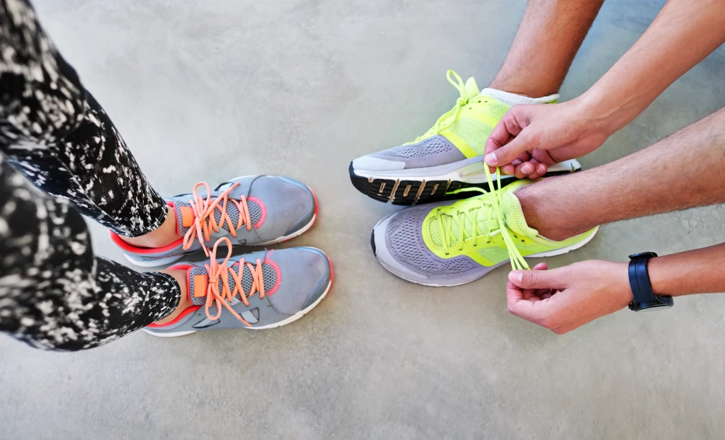 Overhead view of two people in running shoes on concrete, one crouching to lace neon yellow trainers, a fit check that is part of how to choose the right shoes for your feet.