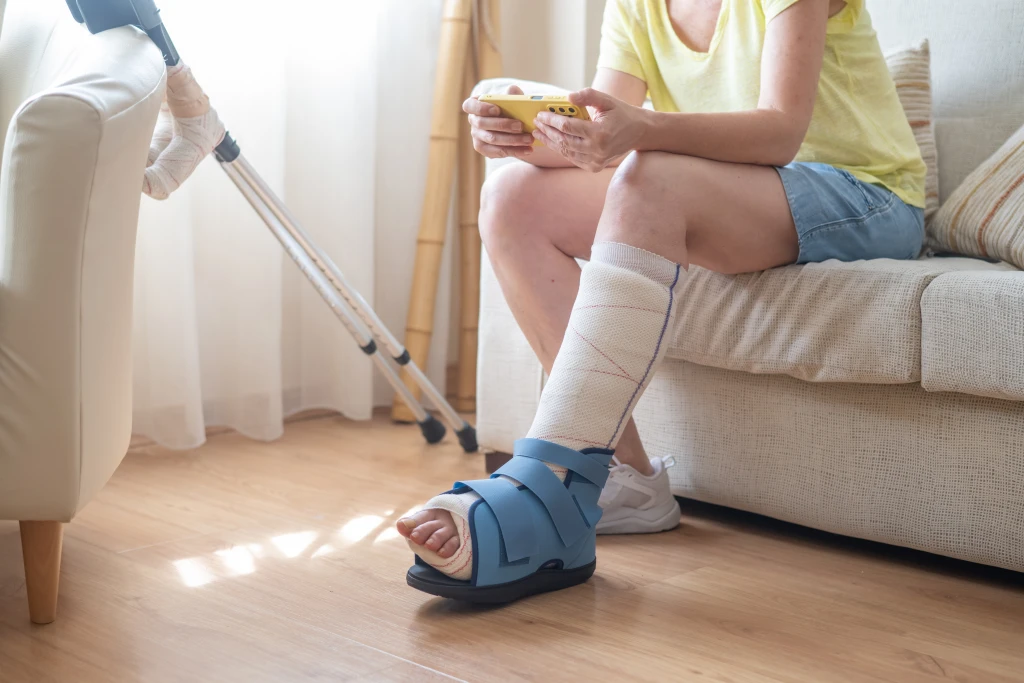 A person sits on a sofa using a phone, one bandaged lower leg in a blue offloading shoe with crutches nearby, the pressure relief behind why offloading matters for a diabetic foot ulcer.
