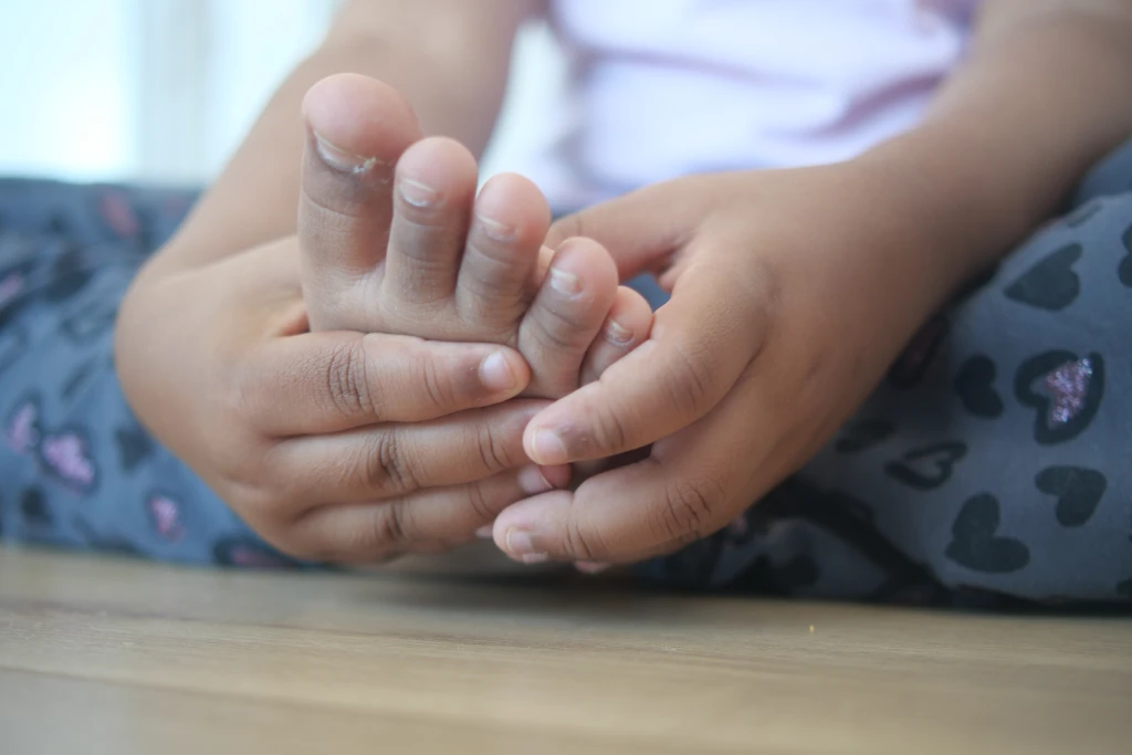 Close-up of a child's hands cradling and squeezing their own bare foot while seated, the self-rubbing that prompts parents to ask how to relieve growing pains in legs.