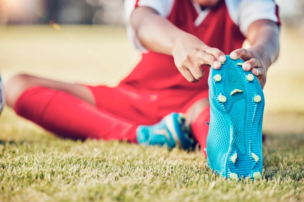 A footballer in a red kit sitting on grass stretching one leg while gripping the studded sole of a blue boot, the studded footwear involved in common football injuries.