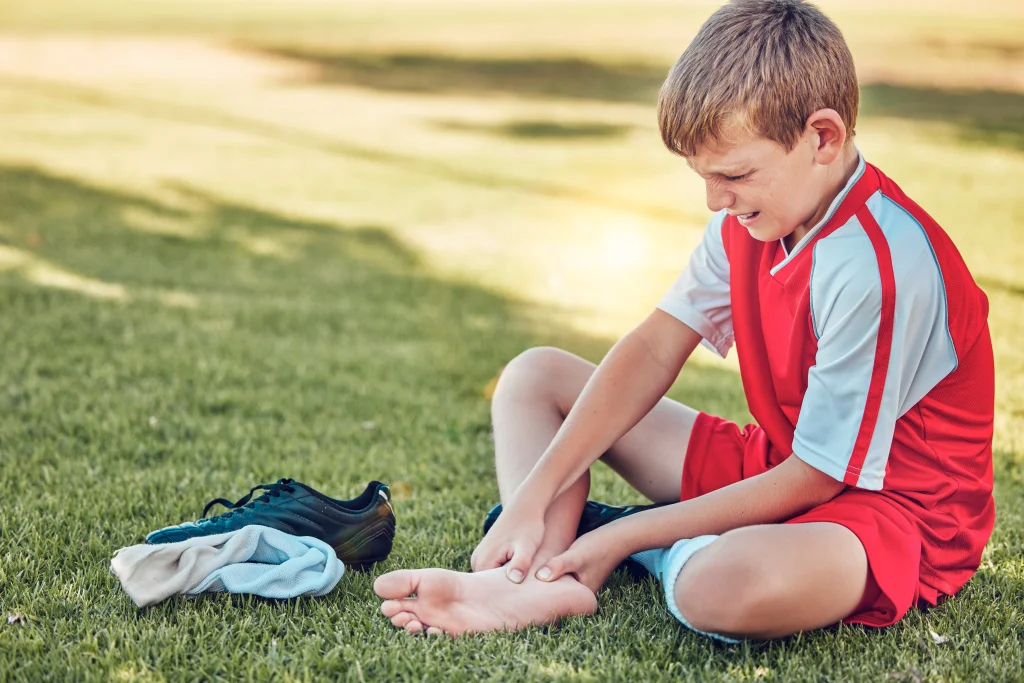 A young footballer in a red kit sits on grass clutching a bare heel beside a discarded boot, the moment that prompts asking how to tell what is causing my heel pain.