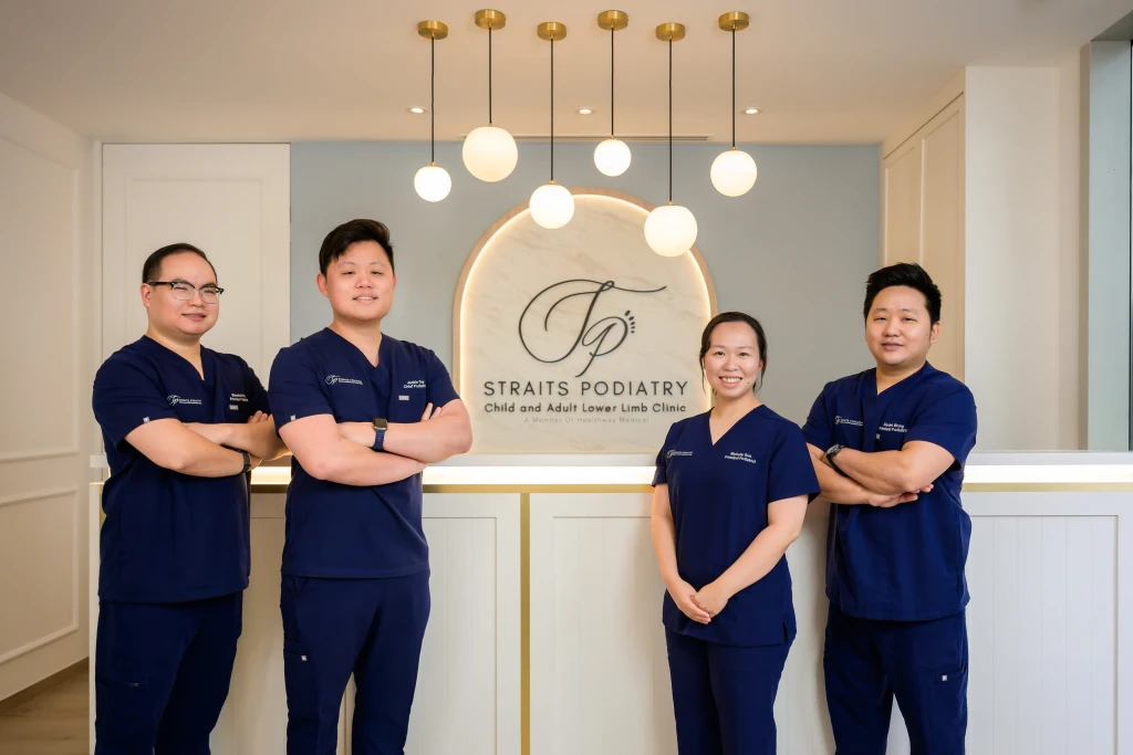 Four podiatrists in navy scrubs stand with folded arms before the clinic reception logo wall, the kind of team patients picture when asking is a podiatrist a doctor