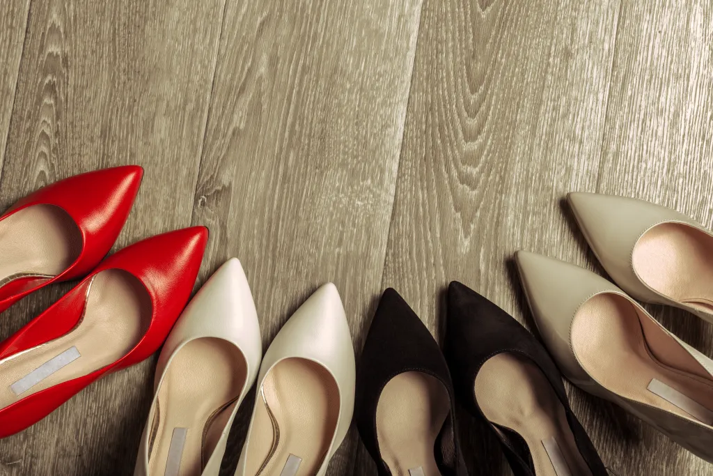 Five pairs of pointed high-heeled shoes lined up on a wood floor, the narrow toe box that can contribute to big toe pain when walking