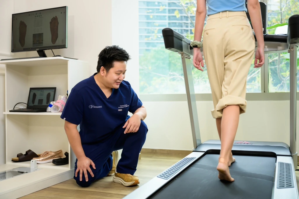 A podiatrist kneeling to watch a patient walk barefoot on a treadmill with foot scan images on the monitor behind, part of what to expect at a podiatry appointment.