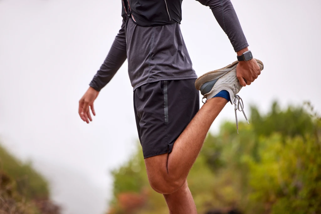 A runner in dark sportswear stands on an outdoor trail pulling one heel up behind to stretch the thigh, a preparation habit used to avoid running injuries.