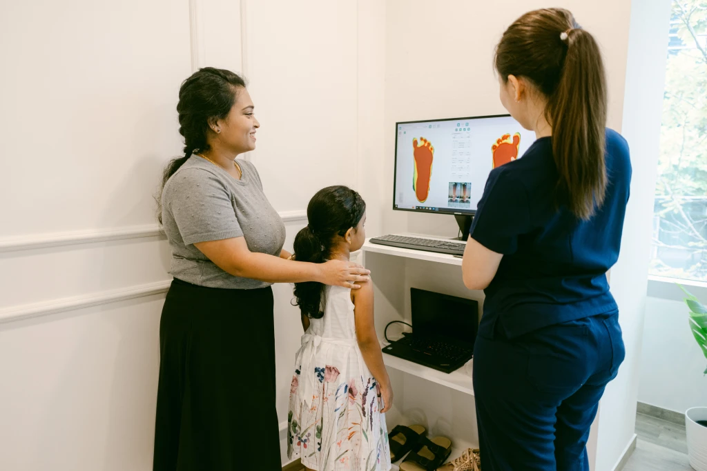 A podiatrist and an adult watching a monitor of colour foot pressure scans with a young child, part of a paediatric assessment for postural issues in children.