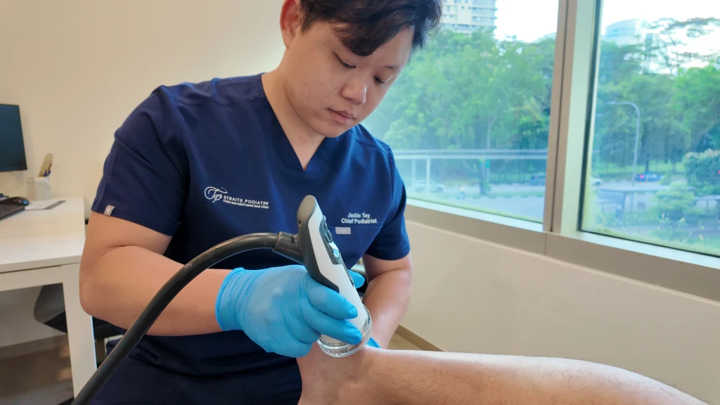 A podiatrist in navy scrubs and blue gloves applies a handheld therapy device to the back of a patient's lower leg, the heel cord region behind achilles tendon function.
