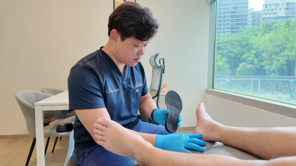A podiatrist in gloves holding a black orthotic insole while assessing a patient's foot on a clinic couch, part of working out what does heel pain mean