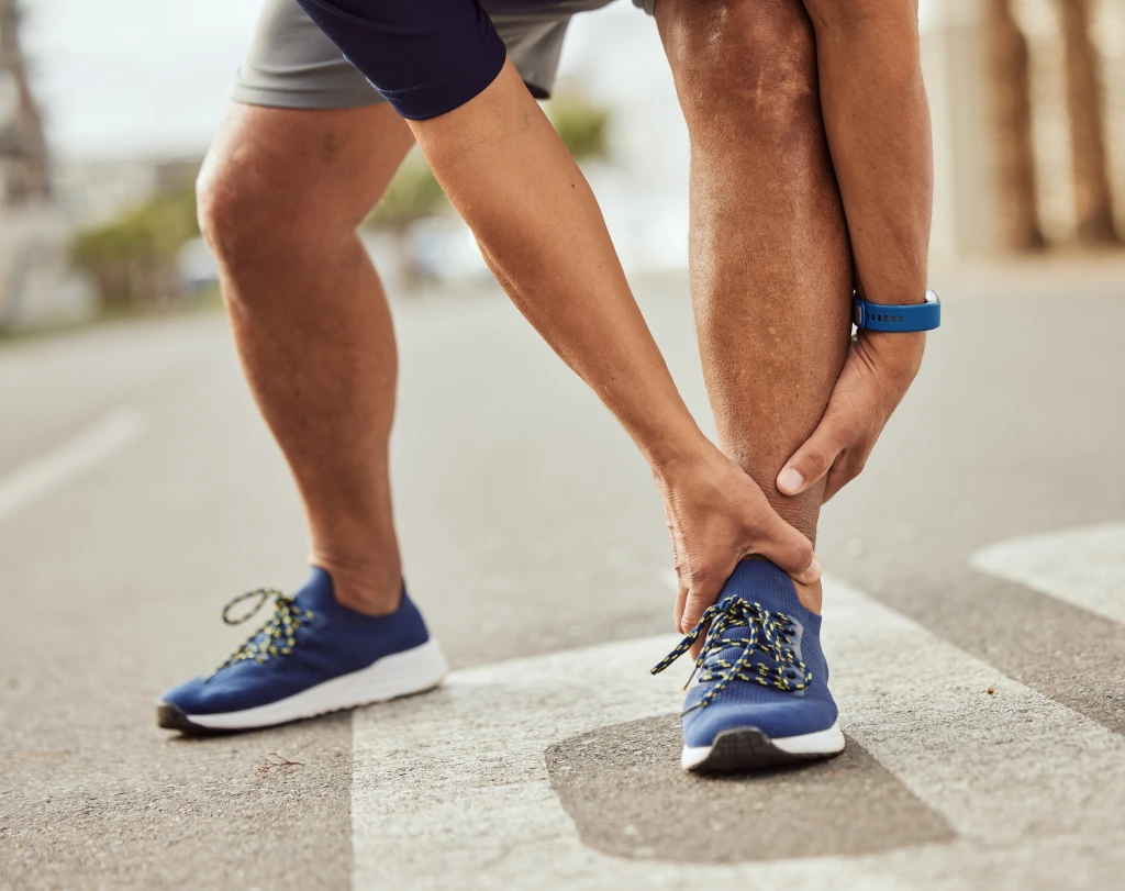 A runner in trainers bending to grip the lower calf and Achilles area on a road, the tendon pain that raises the tendonitis vs tendinosis distinction.