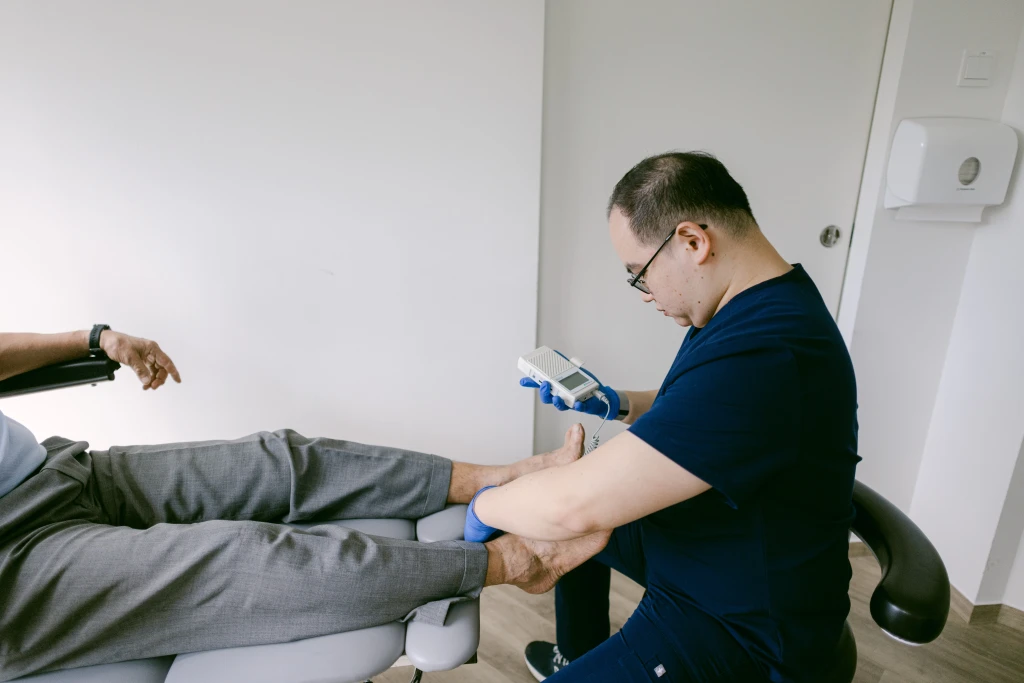 A podiatrist in navy scrubs and gloves holds a handheld Doppler probe to a seated patient's foot to check blood flow, a core step in how to prevent diabetic foot amputation